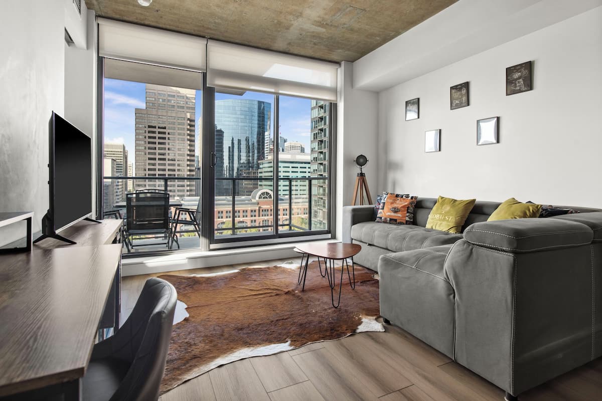 Ein modernes Wohnzimmer mit einem grauen Sofa, einem Holztisch, einem Ledersessel und einem großen Fenster mit Blick auf die Stadt.