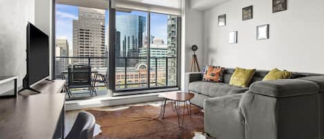 A modern living room with a grey sofa, a wooden coffee table, a leather chair, and a large window offering a city view.