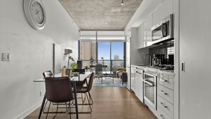 Eine moderne Küche mit weißen Schränken, einer Mikrowelle und einer Kaffeemaschine. Es gibt eine Essecke mit Tisch und Stühlen sowie ein großes Fenster mit Blick auf die Stadt.