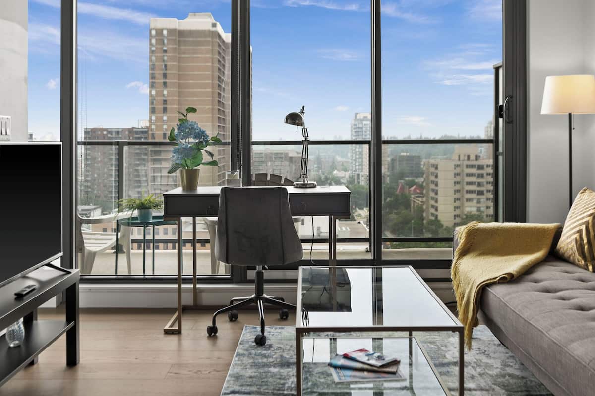 Ein modernes Wohnzimmer mit einem Glastisch, einer Couch, einem Schreibtisch mit Stuhl, einer Zimmerpflanze und Blick auf die Stadt durch große Fenster.