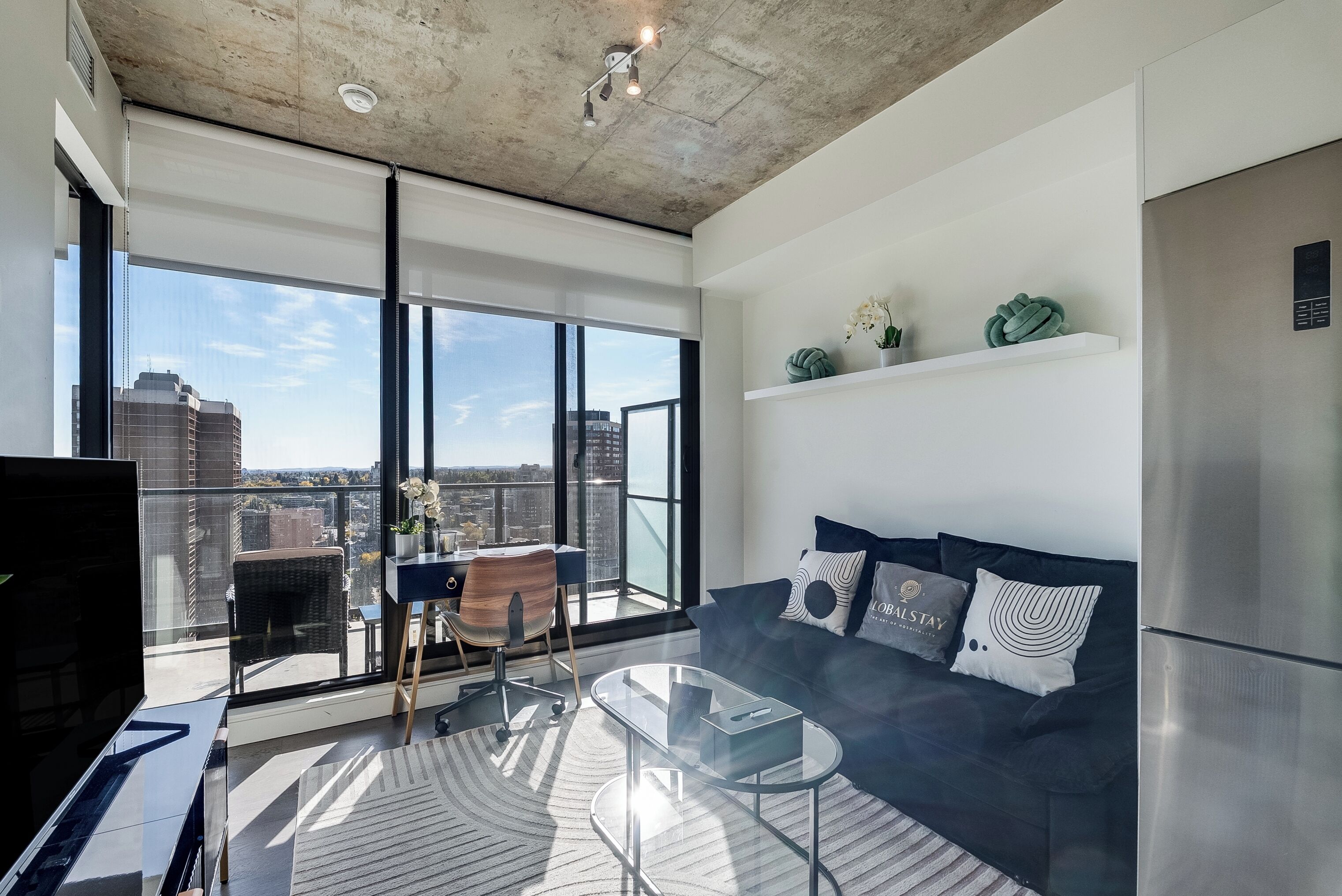 Una sala de estar moderna con una mesa de centro de vidrio, un sofá azul oscuro y vistas a la ciudad a través de amplios ventanales.
