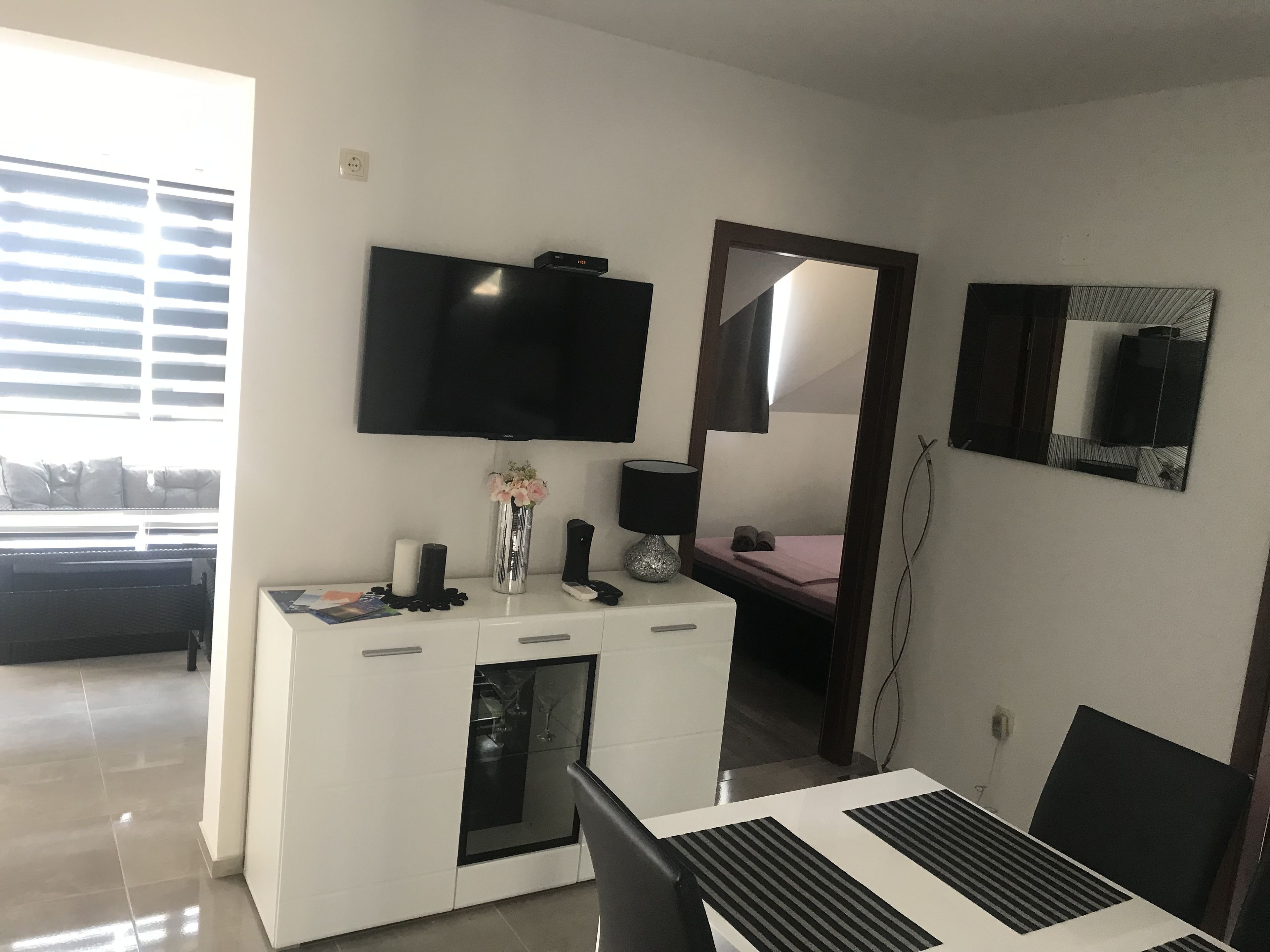 Una sala de estar moderna con un televisor de pantalla plana montado en la pared, un mueble blanco con un jarrón de flores, una mesa de comedor con un camino de mesa blanco y negro, y un espejo grande en la pared.