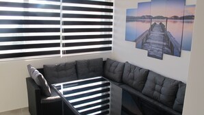 A corner seating area with a cushioned sofa and a small table, a window with blinds, and a wall-mounted picture of a pier. - Comfortable 2 Bedroom Apartment (Mljet)