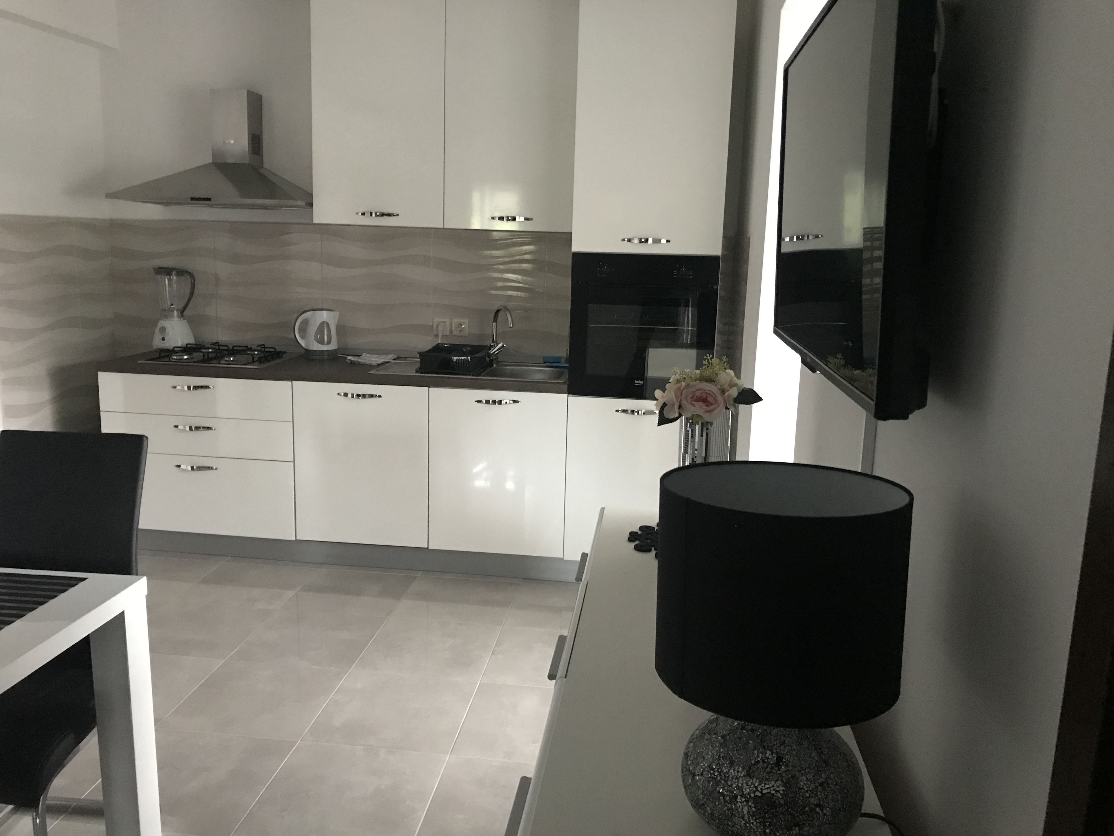 Een moderne keuken met witte kasten, een gasfornuis, een waterkoker en een spoelbak. Er is een eethoek met een tafel en stoelen. Een zwarte lamp staat op een standaard, en er staat een vaas met bloemen op het aanrecht.