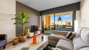 A modern living room with a sofa, coffee table, and TV. There is a large window with a view of buildings and a balcony.