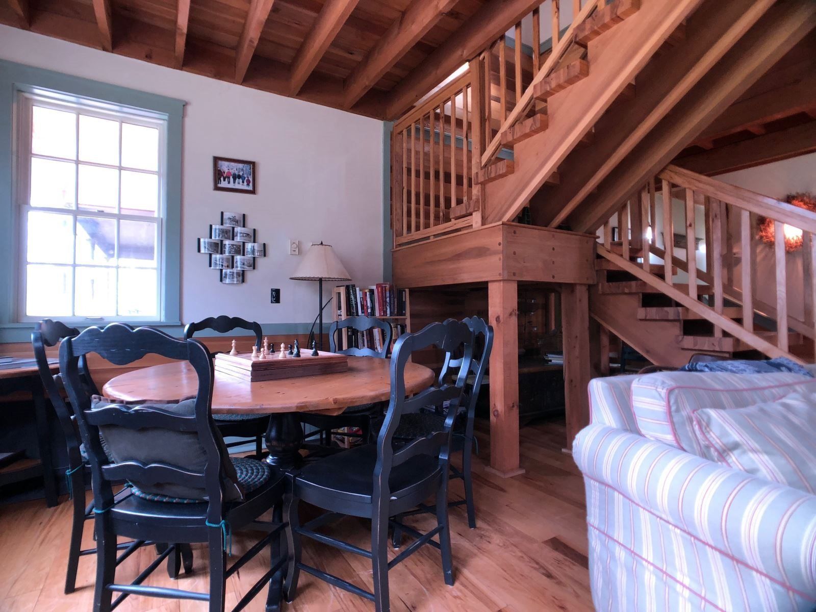 Una mesa de comedor de madera con sillas, una escalera y un sofá en una habitación con vigas de madera en el techo.