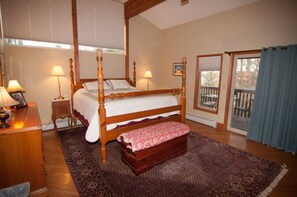 A bedroom with a four-poster bed, a wooden bench, two bedside tables with lamps, a large window with blinds, and a balcony with a view.