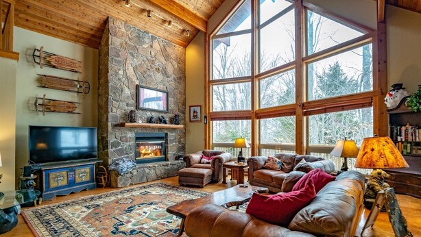 A cozy living room with a stone fireplace, leather sofas, a large TV, and a view of trees through large windows.