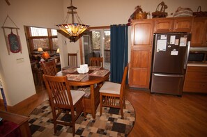 A dining area with a wooden table, chairs, and a patterned rug. There is a chandelier above the table and a refrigerator to the right.