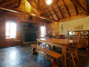 Interior rústico de una cabaña con chimenea de piedra, mesa de comedor de madera, bancos y una zona de cocina con estufa.