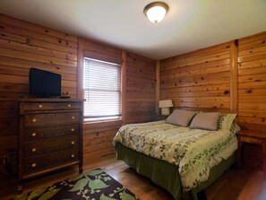 Dormitorio con paneles de madera, una cama, un tocador, una lámpara y un televisor sobre el tocador.