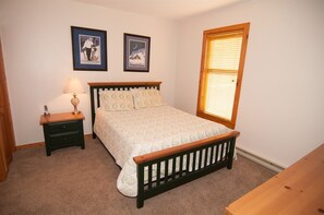 A bedroom with a bed, a nightstand, a lamp, two framed pictures on the wall, and a window with blinds.