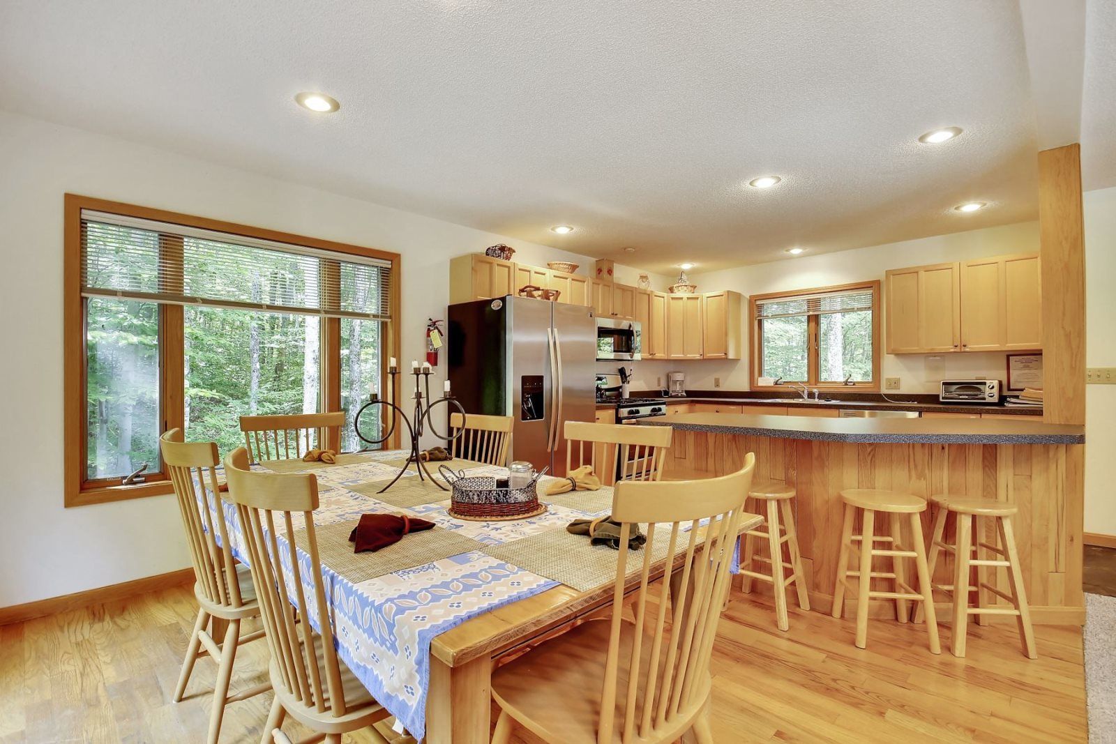 A modern kitchen with wooden cabinets, a central island with stools, a dining table with chairs, and large windows offering a view of the outdoors.