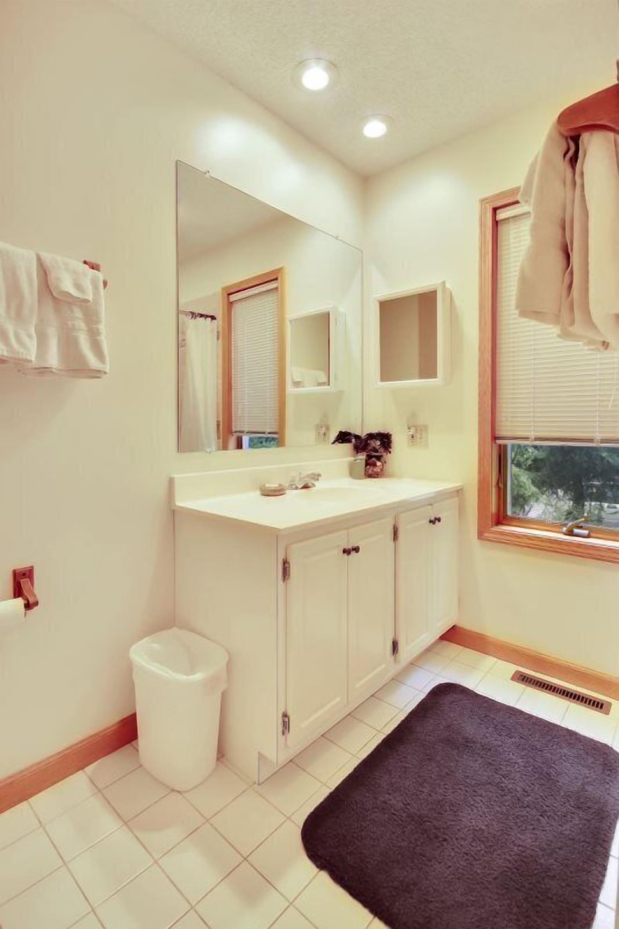 A bathroom with a white sink, mirror, and cabinets. There is a towel rack on the wall and a window with blinds.