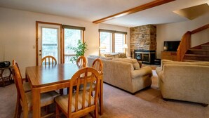 A cozy living room with a stone fireplace, a dining area with wooden furniture, and a staircase leading to an upper level. - Deerfield Village 106 (Davis)
