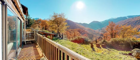 Terraza de madera con barandilla de vidrio que da a un paisaje montañoso con follaje otoñal.