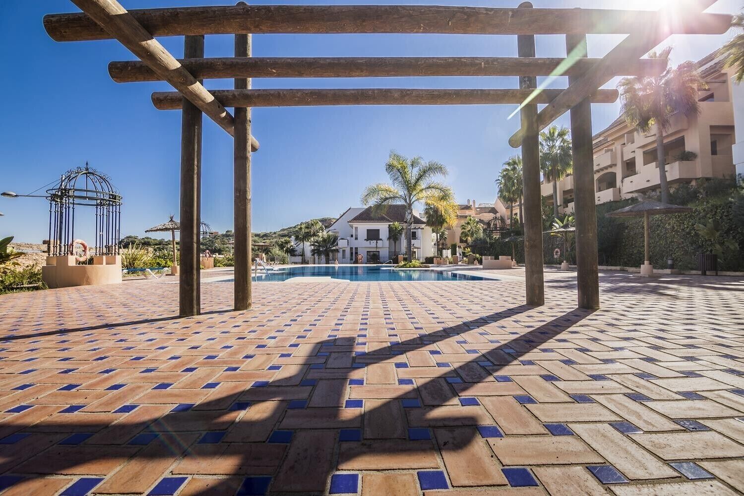 Un espace piscine agrémenté d’une pergola, un ciel d’un bleu limpide et un bâtiment en arrière-plan.