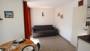 A compact living space with a dining area, a gray sofa, a small kitchen with a refrigerator, and a wall-mounted clock.