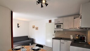 A compact kitchen with a microwave, a small refrigerator, and a sink. There is a dining table set with plates, glasses, and cutlery. A grey sofa is against the wall, and there are two framed pictures on the wall.