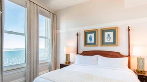 A hotel room with a large bed, two framed pictures on the wall, a nightstand with a lamp, and a view of the sea through the window.