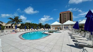 Un espace piscine avec des chaises longues et des parasols, un ciel d’un bleu éclatant et un grand immeuble en arrière-plan.