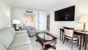 A modern living room with a sofa, a glass coffee table, a dining table with chairs, a TV mounted on the wall, and a door leading to another room. - Marco Beach Ocean Resort 610: Private 1 Bedroom condo; New Tile,TV's (Marco Island)
