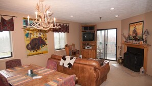A cozy living room with a brown leather sofa, a wooden dining table with colorful placemats, a chandelier, a fireplace, and a television on a wooden stand. - Beaver Ridge 260 (Davis)