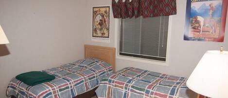 A room with two beds, a window with blinds, a lamp, and two posters on the wall.