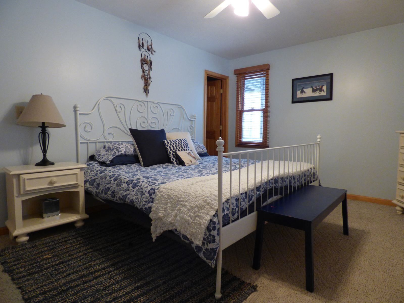 A bedroom with a bed, bedside table, lamp, window, and a framed picture on the wall.