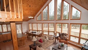 Ein geräumiges Wohnzimmer mit einem großen Holzesstisch, mehreren Stühlen, einer Couch und einem Couchtisch. Große Fenster sorgen für viel natürliches Licht und Ausblick ins Freie.