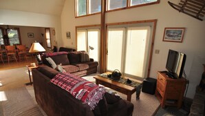 A cozy living room with a brown sofa, a coffee table, and a television on a wooden stand. There are large windows with blinds, a dining area in the background, and a rug on the floor. - Bear`s Den - Northface (Davis)