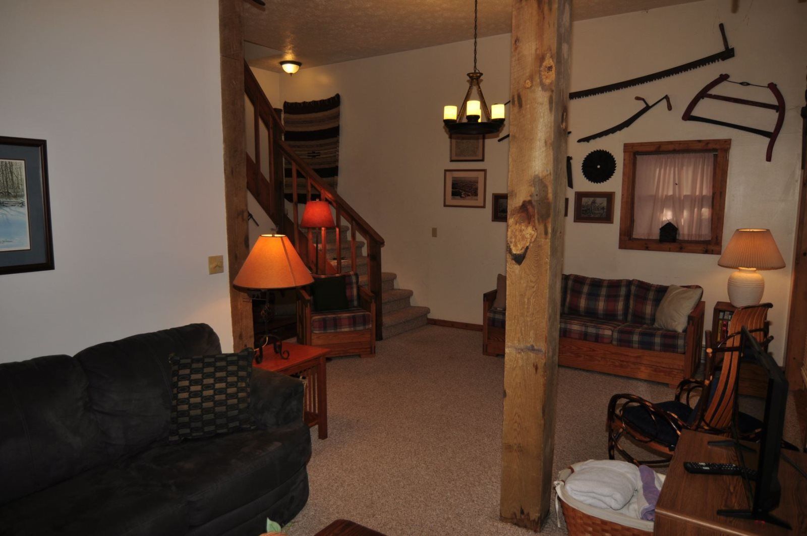 A cozy living room with a dark-colored sofa, a wooden coffee table, a plaid sofa, and a wooden chair. A staircase with a wooden railing leads to another level.