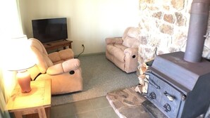 Ett mysigt vardagsrum med en steneldstad, två beige fåtöljer, ett träbord och en tv som står på ett stativ.