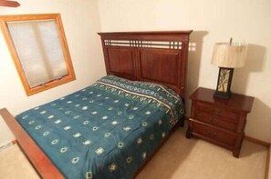 A bedroom with a wooden bed, a patterned bedspread, a nightstand with a lamp, and a window with blinds.