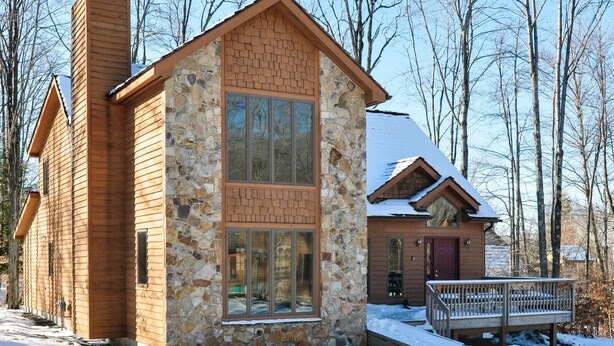 A two-story log cabin with a stone facade and large windows, surrounded by a snowy landscape and trees. - Almost Heaven (Davis)