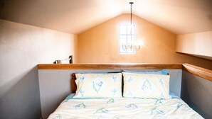 Une chambre avec un plafond en pente, une fenêtre avec un luminaire et un lit recouvert d’une literie à motifs floraux.
