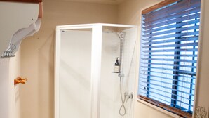 Une salle de bain avec un carrelage à motifs géométriques, des toilettes blanches avec un abattant en bois, un espace douche et une fenêtre avec des stores.