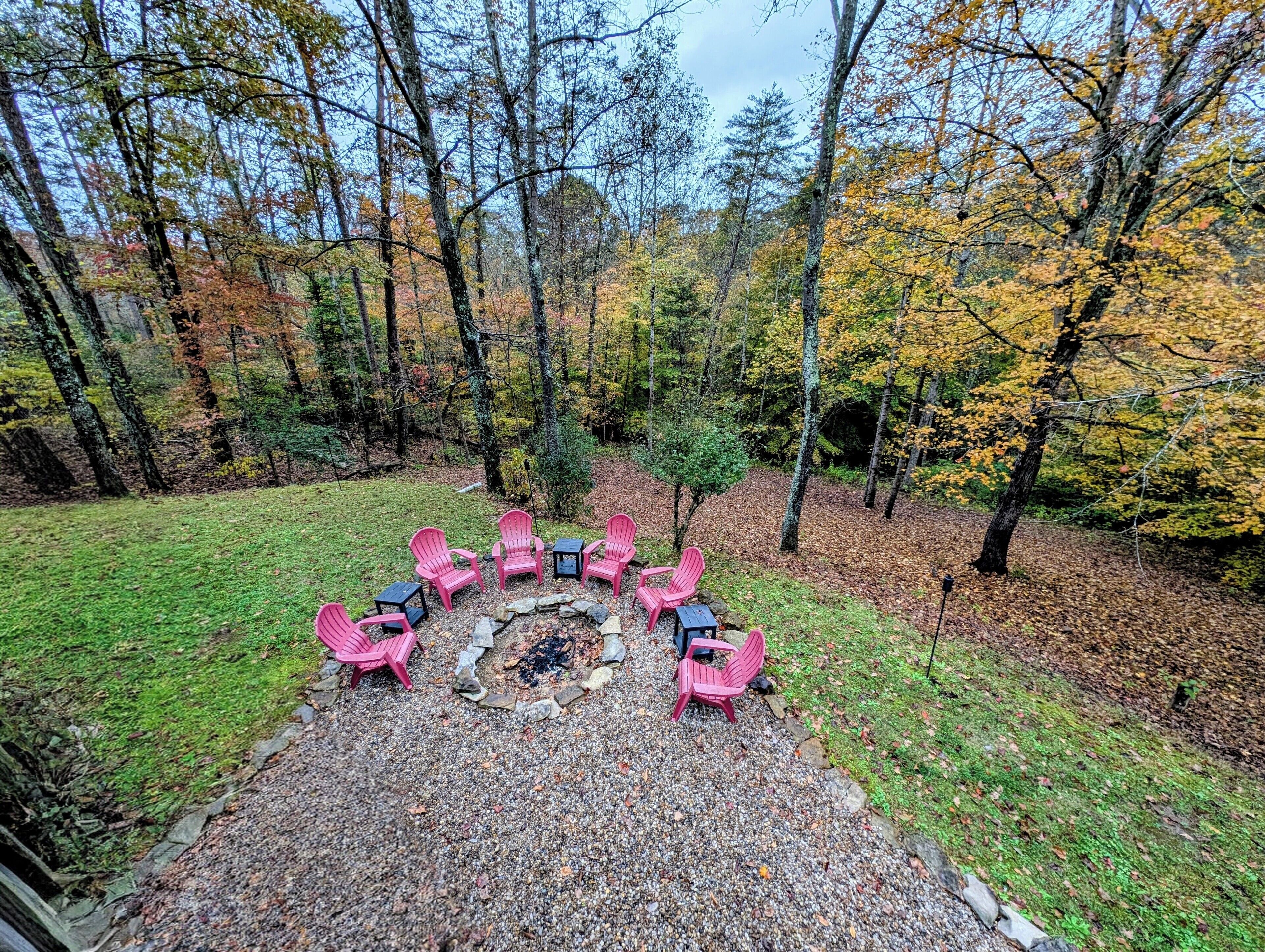 A circular seating area with six pink Adirondack chairs around a central fire pit, surrounded by a gravel path and a grassy lawn, set in a forested area with autumn-colored trees.