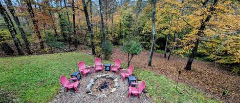 A circular seating area with six pink Adirondack chairs around a central fire pit, surrounded by a gravel path and a grassy lawn, set in a forested area with autumn-colored trees.