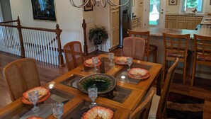 A dining area with a wooden table set for four, surrounded by wicker chairs. A chandelier hangs above, and a large window provides natural light.