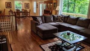 A living room with a sectional sofa, a glass-top coffee table, wooden flooring, a dining area with a table and chairs, and a view of greenery through large windows.