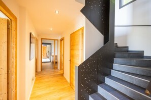 Um interior moderno com um corredor de madeira que leva a uma escadaria com degraus pretos e uma porta de madeira no final.