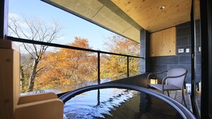 Un baño moderno con una bañera de hidromasaje con mampara de vidrio, techo de madera y vistas a árboles otoñales.