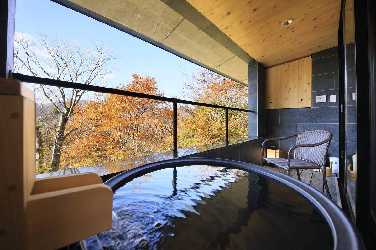 Un bagno moderno con vasca idromassaggio in vetro, soffitto in legno e vista sugli alberi autunnali.