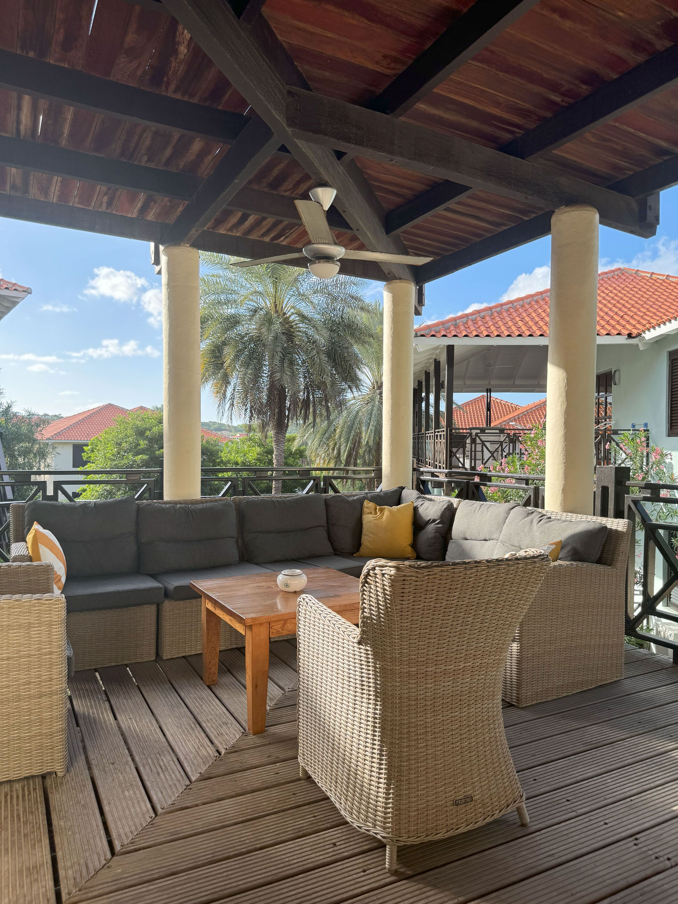 A covered outdoor seating area with a wooden table, wicker furniture, and a view of a residential area with red-tiled roofs and palm trees.
