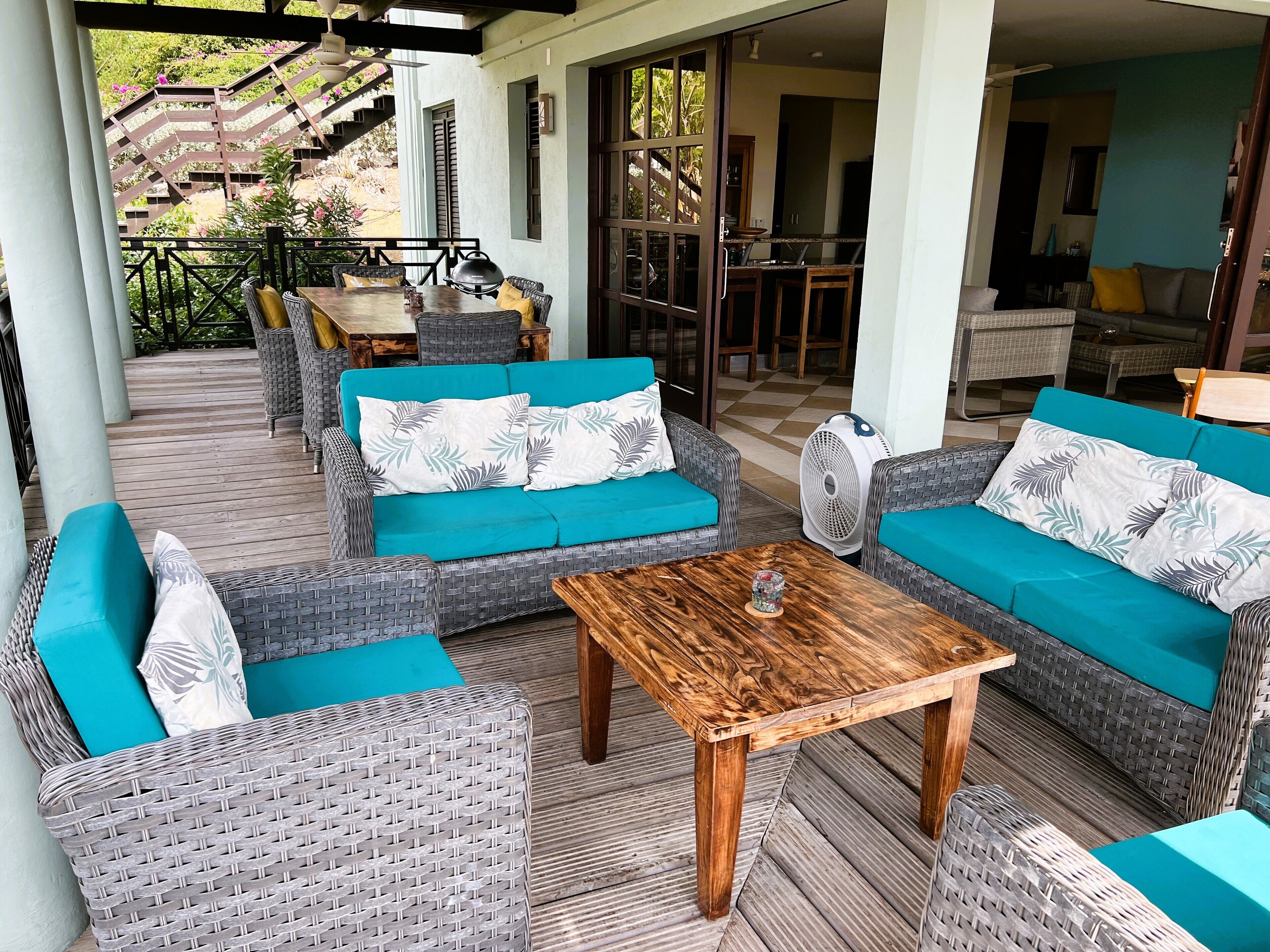 Ein überdachter Außenbereich mit Korbmöbeln und blaumetallenen Kissen, einem Holztisch und Blick auf den Garten und die Küche.