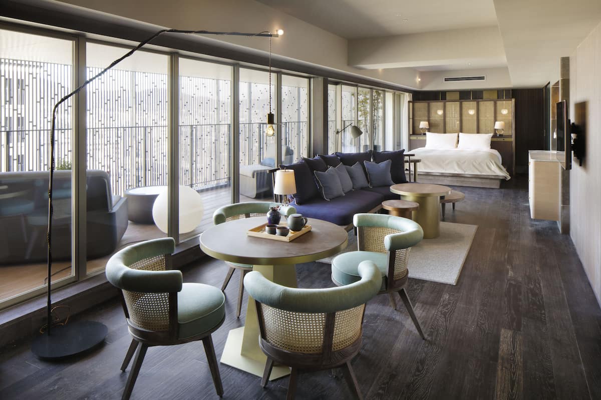 Ein modernes Hotelzimmer mit einem großen Bett, einer Couch, einem Esstisch mit Stühlen und Blick durch große Fenster.