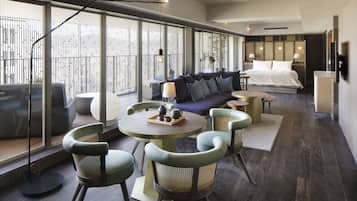 A modern hotel room with a large bed, a sofa, a dining table with chairs, and a view through large windows.