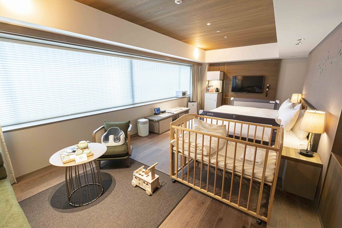 Ein Hotelzimmer mit Bett, Schreibtisch, Stuhl, Babybett, Fernseher und einem Fenster mit Jalousien.