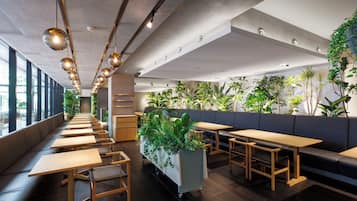 Un restaurante moderno con largas mesas y bancos de madera, un macetero central con follaje y un techo alto con lámparas colgantes.